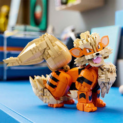 A toy building-block animal character resembling a fox or tiger, assembled from orange and beige bricks, standing on a blue surface with a blurred indoor background.