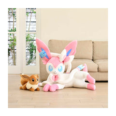 Two plush toys, one small and one large, on a wooden floor with a beige couch and window in the background.