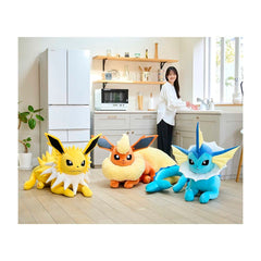 Three large plush Pokémon toys are displayed on a light wood floor in a bright kitchen setting, with a woman standing in the background near the cabinets and refrigerator.