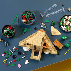 A LEGO building set arranged on a dark blue-gray surface, showing a partially assembled tan house with bowls of loose bricks, clear connectors, small accessories, and a mini-doll figure.