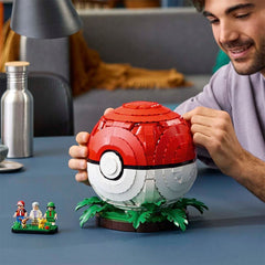 A man holds a red and white spherical Pokéball-style decorative object on a green leaf base at a table, with small minifigures nearby and a lamp and bottle in the background.